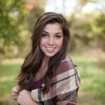 A woman smiling outdoors with a cozy plaid shawl, surrounded by nature in a sunny park.